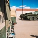 The image shows military vehicles in what appears to be a base or staging area under a clear blue sky. In the foreground, a close-up of a military vehicle panel displays stenciled markings reading '4ID10N' in black lettering on a two-tone green and tan camouflage surface. In the background, a green 8-wheeled armored vehicle, consistent with a Stryker-type APC, is parked near a building.