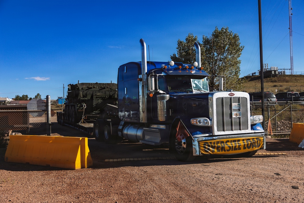 A blue Peterbilt semi-truck with an 'OVERSIZE LOAD' banner on its front bumper is parked or moving through what appears to be a secured facility entrance with yellow concrete barriers. The truck is hauling a flatbed trailer carrying large, heavy green military or industrial equipment secured with chains. The setting appears to be a rural or semi-industrial area under a clear blue sky.