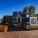 A blue Peterbilt semi-truck with an 'OVERSIZE LOAD' banner on its front bumper is parked or moving through what appears to be a secured facility entrance with yellow concrete barriers. The truck is hauling a flatbed trailer carrying large, heavy green military or industrial equipment secured with chains. The setting appears to be a rural or semi-industrial area under a clear blue sky.