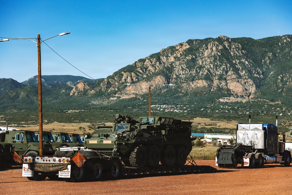 A large military armored wheeled vehicle, consistent with an M1126 Stryker Infantry Carrier Vehicle, is loaded on a flatbed semi-trailer in a dirt lot. The scene is set against a dramatic Rocky Mountain backdrop with clear blue skies, suggesting a location near Colorado Springs, Colorado. Additional military trucks are visible in the background, and a commercial semi-truck cab is attached to the flatbed.