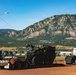 A large military armored wheeled vehicle, consistent with an M1126 Stryker Infantry Carrier Vehicle, is loaded on a flatbed semi-trailer in a dirt lot. The scene is set against a dramatic Rocky Mountain backdrop with clear blue skies, suggesting a location near Colorado Springs, Colorado. Additional military trucks are visible in the background, and a commercial semi-truck cab is attached to the flatbed.