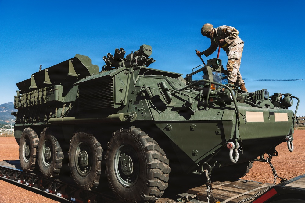 A soldier in camouflage uniform and helmet is working on top of a large olive-green 8-wheeled armored fighting vehicle, identified as a Stryker variant, loaded on a flatbed transport trailer. The vehicle is equipped with multiple weapon systems and smoke grenade launchers mounted on its hull. The scene appears to be at a military staging or transport area under clear blue skies.