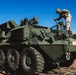 A soldier in camouflage uniform and helmet is working on top of a large olive-green 8-wheeled armored fighting vehicle, identified as a Stryker variant, loaded on a flatbed transport trailer. The vehicle is equipped with multiple weapon systems and smoke grenade launchers mounted on its hull. The scene appears to be at a military staging or transport area under clear blue skies.