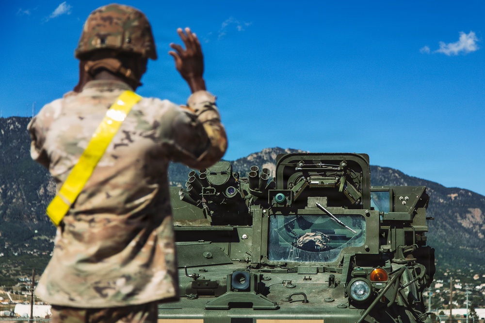 A soldier in MultiCam/OCP camouflage uniform with a yellow reflective safety sash is directing a military armored vehicle, likely a Stryker variant, using hand signals. The scene appears to take place at a military installation with mountainous terrain visible in the background, consistent with locations such as Fort Carson, Colorado. A second soldier is visible through the vehicle's windshield, receiving the directional guidance.