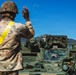 A soldier in MultiCam/OCP camouflage uniform with a yellow reflective safety sash is directing a military armored vehicle, likely a Stryker variant, using hand signals. The scene appears to take place at a military installation with mountainous terrain visible in the background, consistent with locations such as Fort Carson, Colorado. A second soldier is visible through the vehicle's windshield, receiving the directional guidance.