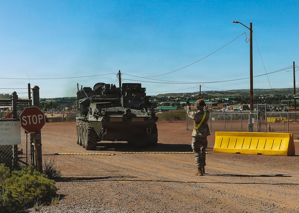 A military soldier in camouflage uniform with a yellow safety vest is directing a large wheeled armored vehicle (consistent with a Stryker-type APC) through a checkpoint or gate entry point. The scene appears to be on a military installation, with security barriers, a stop sign, and chain-link fencing visible. The vehicle is emitting exhaust smoke as it maneuvers through the controlled access point.