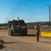 A military soldier in camouflage uniform with a yellow safety vest is directing a large wheeled armored vehicle (consistent with a Stryker-type APC) through a checkpoint or gate entry point. The scene appears to be on a military installation, with security barriers, a stop sign, and chain-link fencing visible. The vehicle is emitting exhaust smoke as it maneuvers through the controlled access point.