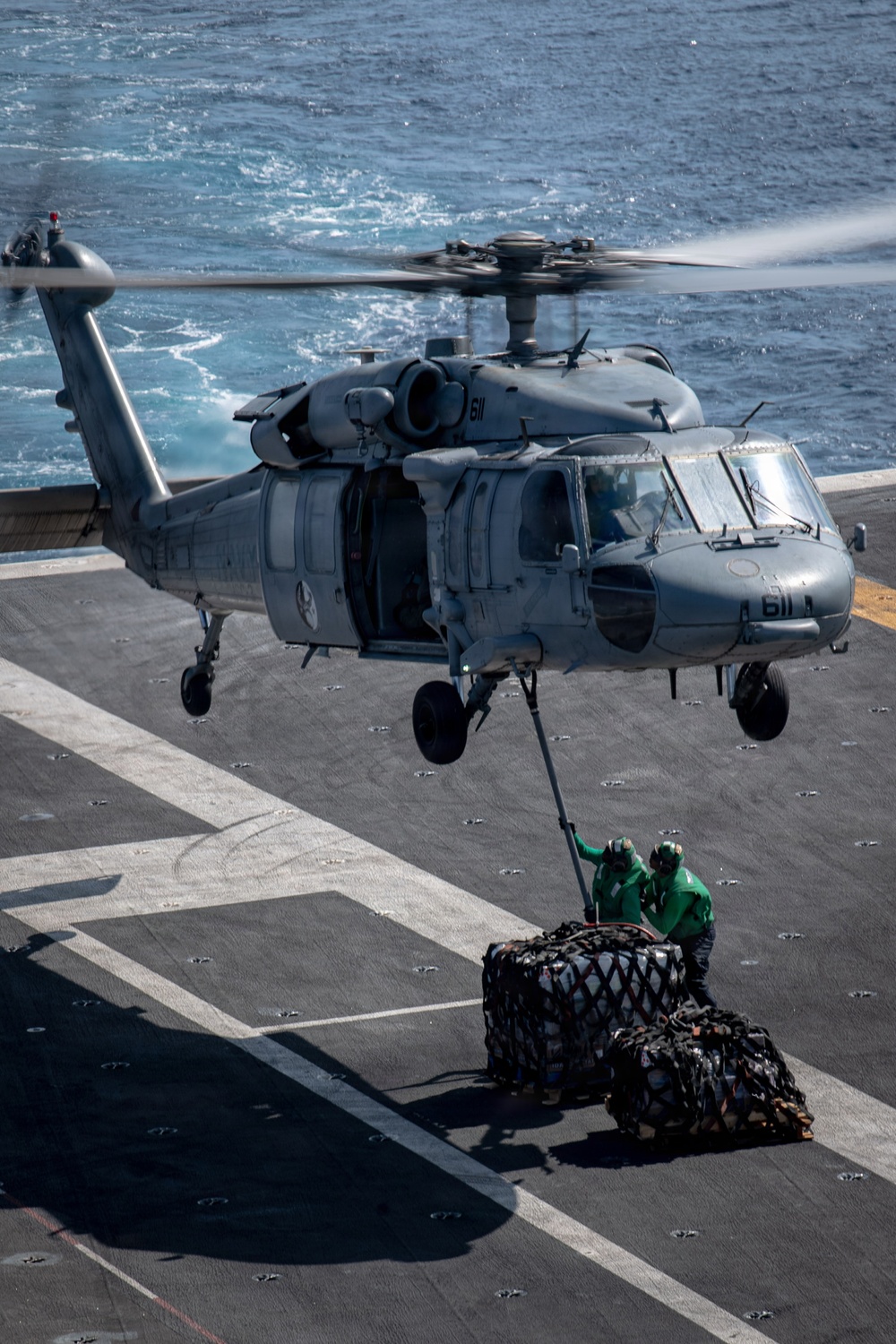 A U.S. Navy MH-60S Seahawk helicopter (bureau number 611) hovers over a ship's flight deck performing a vertical replenishment (VERTREP) operation at sea. Two deck crew members dressed in green jerseys and helmets are on deck attaching or detaching a cargo net loaded with supplies to the helicopter's external hoist. The ocean is visible in the background, indicating the operation is conducted underway.