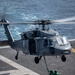 A U.S. Navy MH-60S Seahawk helicopter (bureau number 611) hovers over a ship's flight deck performing a vertical replenishment (VERTREP) operation at sea. Two deck crew members dressed in green jerseys and helmets are on deck attaching or detaching a cargo net loaded with supplies to the helicopter's external hoist. The ocean is visible in the background, indicating the operation is conducted underway.
