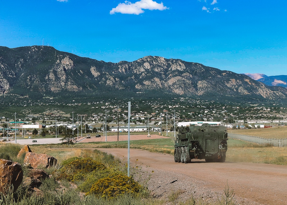 A military wheeled armored vehicle, consistent with a Stryker-type 8x8 armored personnel carrier, travels along a dirt road on what appears to be a U.S. military installation. The backdrop features a dramatic mountain range, a small town or base community, and open terrain under a clear blue sky. The setting is consistent with Fort Carson, Colorado, given the distinctive Cheyenne Mountain range visible in the background.