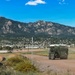A military wheeled armored vehicle, consistent with a Stryker-type 8x8 armored personnel carrier, travels along a dirt road on what appears to be a U.S. military installation. The backdrop features a dramatic mountain range, a small town or base community, and open terrain under a clear blue sky. The setting is consistent with Fort Carson, Colorado, given the distinctive Cheyenne Mountain range visible in the background.