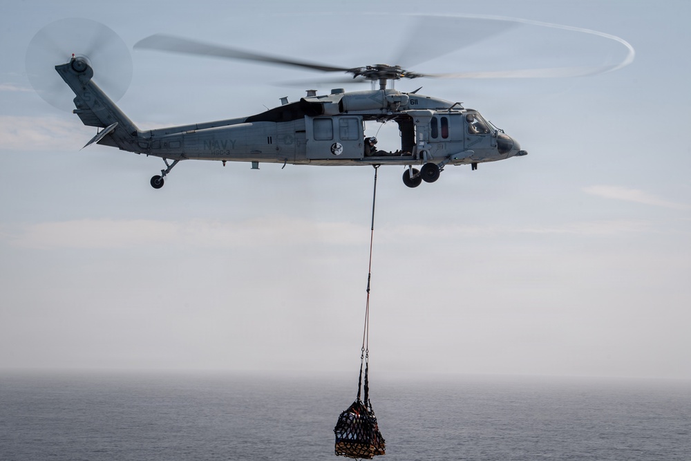 A U.S. Navy helicopter, marked with the number 611 and 'NAVY' on the fuselage, is hovering over open ocean while conducting a vertical replenishment (VERTREP) operation. The helicopter is carrying a cargo net loaded with supplies suspended beneath it via a long sling line. The image captures a military logistics operation at sea under overcast skies.
