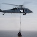 A U.S. Navy helicopter, marked with the number 611 and 'NAVY' on the fuselage, is hovering over open ocean while conducting a vertical replenishment (VERTREP) operation. The helicopter is carrying a cargo net loaded with supplies suspended beneath it via a long sling line. The image captures a military logistics operation at sea under overcast skies.