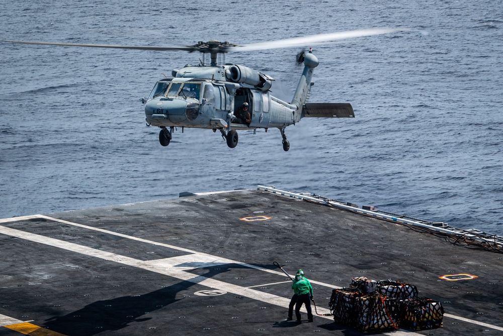 A U.S. Navy helicopter, identified by the number '611' on its nose, hovers over a ship's flight deck during what appears to be a vertical replenishment (VERTREP) or cargo transfer operation at sea. A deck crew member in a green jersey stands on the flight deck holding a guide line or hook pole, with netted cargo pallets visible nearby. The ocean is visible in the background, indicating the vessel is underway.