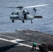 A U.S. Navy helicopter, identified by the number '611' on its nose, hovers over a ship's flight deck during what appears to be a vertical replenishment (VERTREP) or cargo transfer operation at sea. A deck crew member in a green jersey stands on the flight deck holding a guide line or hook pole, with netted cargo pallets visible nearby. The ocean is visible in the background, indicating the vessel is underway.