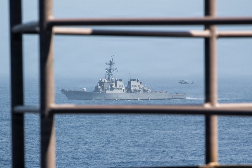 A naval warship, consistent with a U.S. Navy Arleigh Burke-class guided-missile destroyer, is photographed through a metal grid or railing structure in the foreground. The vessel is underway on open blue water, accompanied by a military helicopter visible to its starboard side. The framing through the metallic structure creates a layered compositional effect.