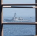 A naval warship, consistent with a U.S. Navy Arleigh Burke-class guided-missile destroyer, is photographed through a metal grid or railing structure in the foreground. The vessel is underway on open blue water, accompanied by a military helicopter visible to its starboard side. The framing through the metallic structure creates a layered compositional effect.