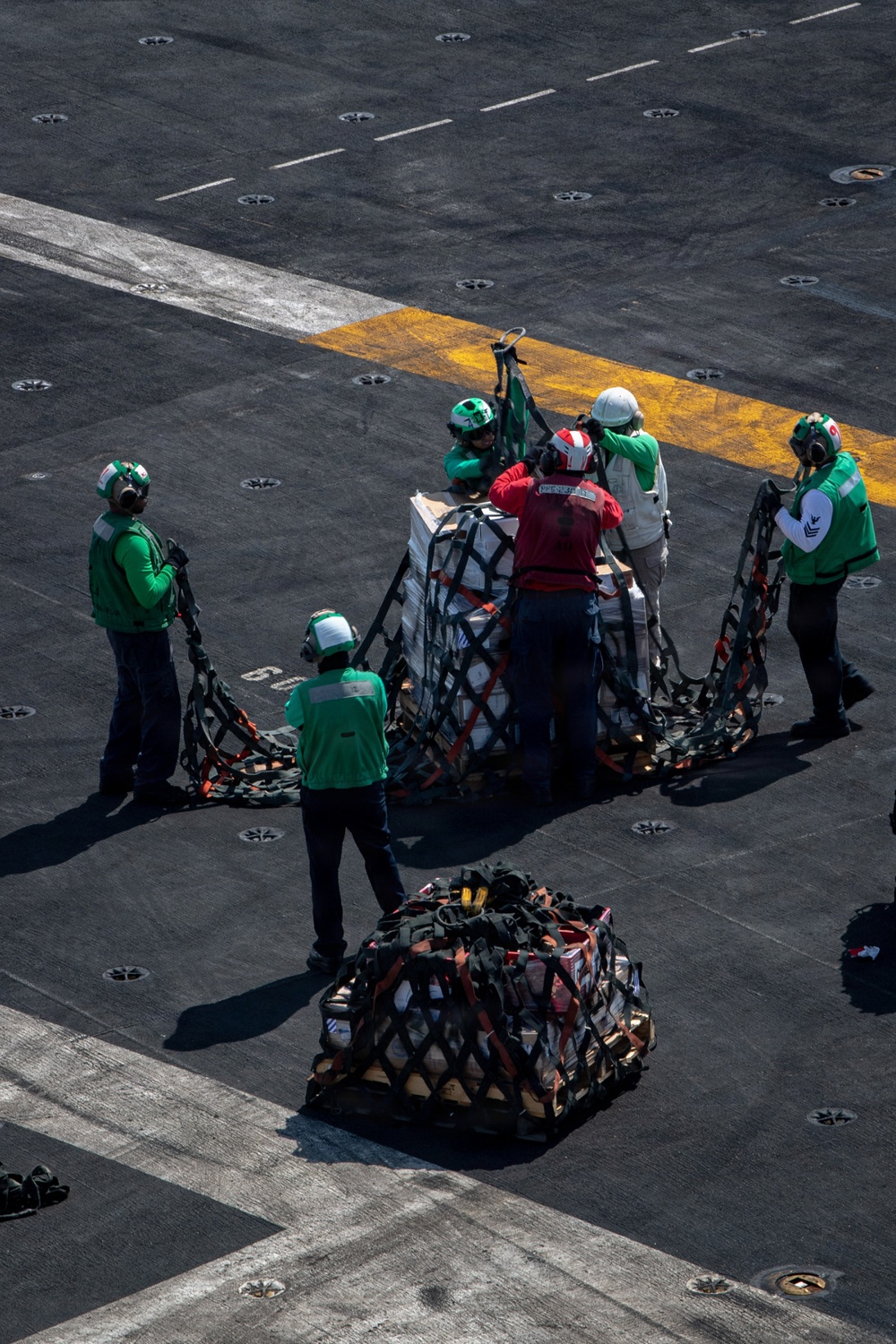 The image shows a group of naval personnel on what appears to be an aircraft carrier flight deck conducting a vertical replenishment (VERTREP) or connected replenishment (CONREP) operation. Crew members in green jerseys and helmets are handling cargo pallets secured with cargo nets and orange straps. A second netted cargo pallet is visible in the foreground, already delivered to the deck.