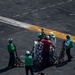 The image shows a group of naval personnel on what appears to be an aircraft carrier flight deck conducting a vertical replenishment (VERTREP) or connected replenishment (CONREP) operation. Crew members in green jerseys and helmets are handling cargo pallets secured with cargo nets and orange straps. A second netted cargo pallet is visible in the foreground, already delivered to the deck.