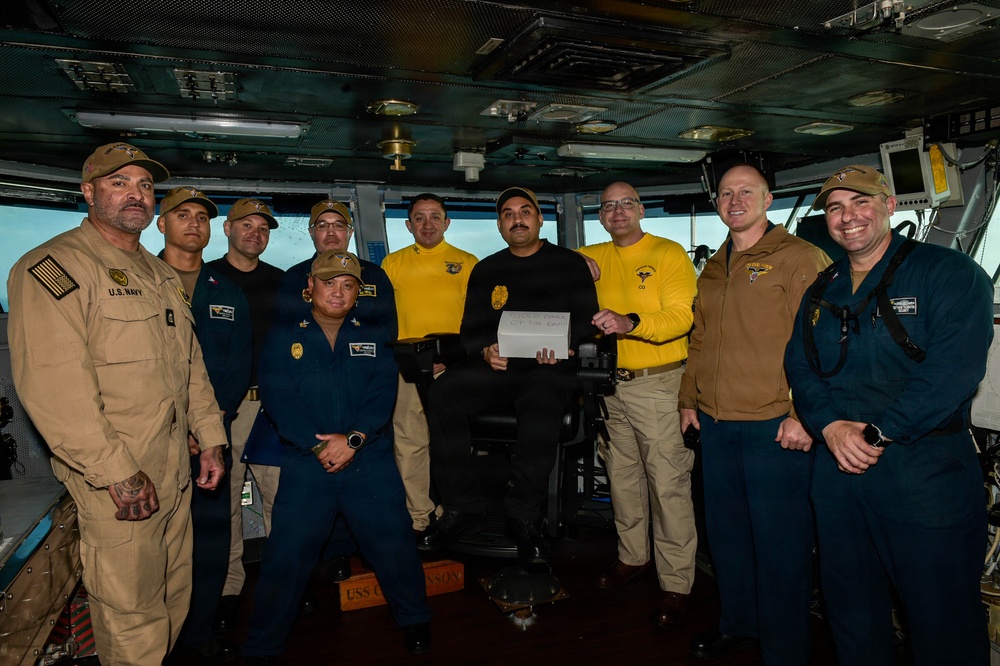 A group of approximately ten U.S. Navy personnel are posed together on what appears to be the bridge of a naval vessel, identifiable by a partially visible 'USS C...' placard on the floor. One central individual in a black shirt is holding a white card or certificate, suggesting a recognition or award ceremony. Personnel are wearing a mix of Navy working uniforms including tan/khaki, blue, and yellow-accented shirts.