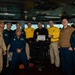 A group of approximately ten U.S. Navy personnel are posed together on what appears to be the bridge of a naval vessel, identifiable by a partially visible 'USS C...' placard on the floor. One central individual in a black shirt is holding a white card or certificate, suggesting a recognition or award ceremony. Personnel are wearing a mix of Navy working uniforms including tan/khaki, blue, and yellow-accented shirts.