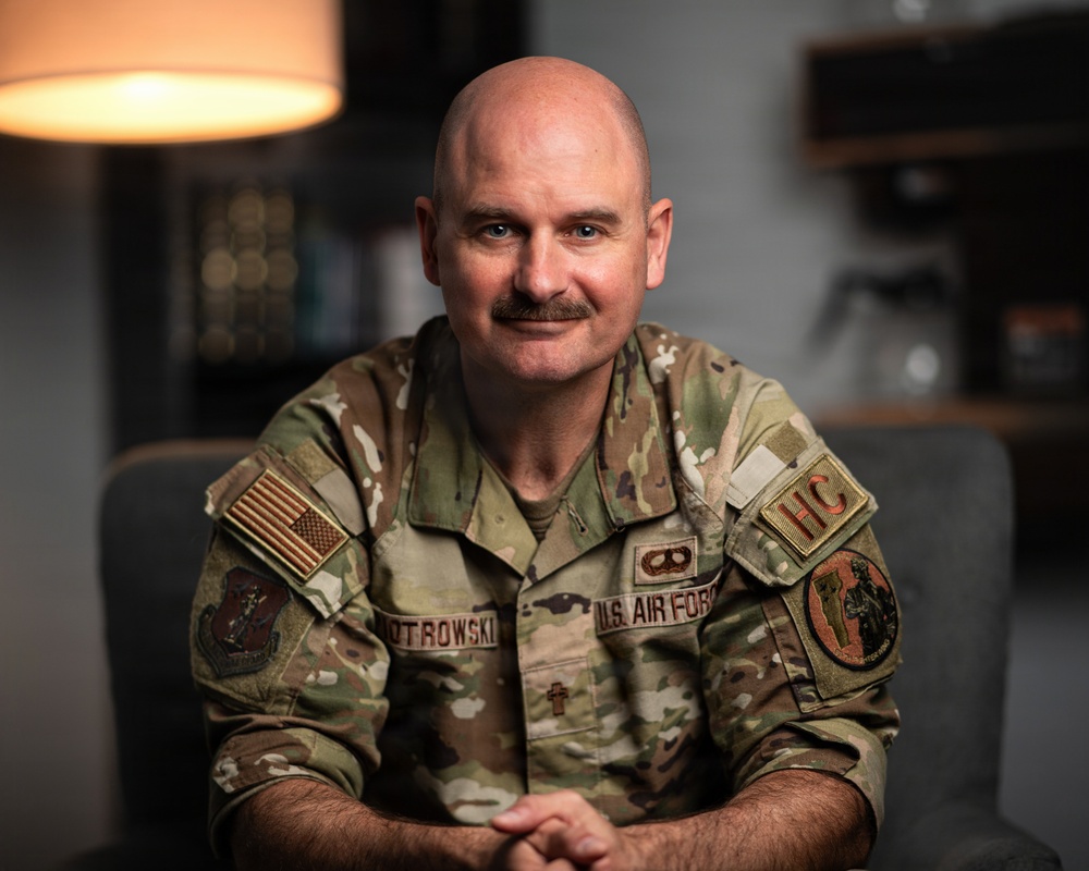 A U.S. Air Force chaplain in OCP (Operational Camouflage Pattern) uniform is seated and posing for a portrait-style photograph. The name tape reads 'PIOTROWSKI' and the uniform displays an 'HC' (Head Chaplain) tab along with a cross insignia, indicating a military chaplain role. The setting appears to be an indoor office or lounge environment with warm ambient lighting.