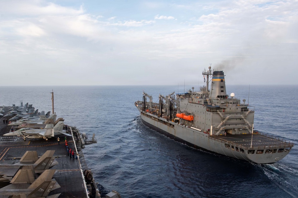 The image shows a replenishment-at-sea (RAS) operation between what appears to be a US Navy aircraft carrier (visible on the left with parked fighter jets on the flight deck) and a naval auxiliary replenishment vessel on the right. Both ships are underway in open ocean, positioned in close parallel formation typical of underway replenishment operations. Personnel in colored jerseys are visible on the carrier's flight deck.