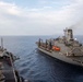 The image shows a replenishment-at-sea (RAS) operation between what appears to be a US Navy aircraft carrier (visible on the left with parked fighter jets on the flight deck) and a naval auxiliary replenishment vessel on the right. Both ships are underway in open ocean, positioned in close parallel formation typical of underway replenishment operations. Personnel in colored jerseys are visible on the carrier's flight deck.