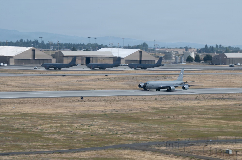 A military air base with a large gray aircraft, likely a KC-135 Stratotanker, taxiing on the runway in the foreground. Multiple similar aircraft are parked on the ramp in the background near large tan-colored hangars. The setting appears to be a U.S. Air Force installation with dry, grassy terrain and mountains visible in the hazy distance.