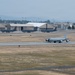A military air base with a large gray aircraft, likely a KC-135 Stratotanker, taxiing on the runway in the foreground. Multiple similar aircraft are parked on the ramp in the background near large tan-colored hangars. The setting appears to be a U.S. Air Force installation with dry, grassy terrain and mountains visible in the hazy distance.
