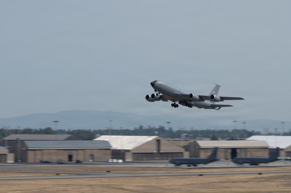 A large military jet aircraft, consistent with a KC-135 Stratotanker, is captured during takeoff from a military airfield. The landing gear is still extended as the aircraft climbs steeply into a hazy sky. Military hangars and additional aircraft are visible on the flight line in the background.