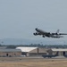 A large military jet aircraft, consistent with a KC-135 Stratotanker, is captured during takeoff from a military airfield. The landing gear is still extended as the aircraft climbs steeply into a hazy sky. Military hangars and additional aircraft are visible on the flight line in the background.