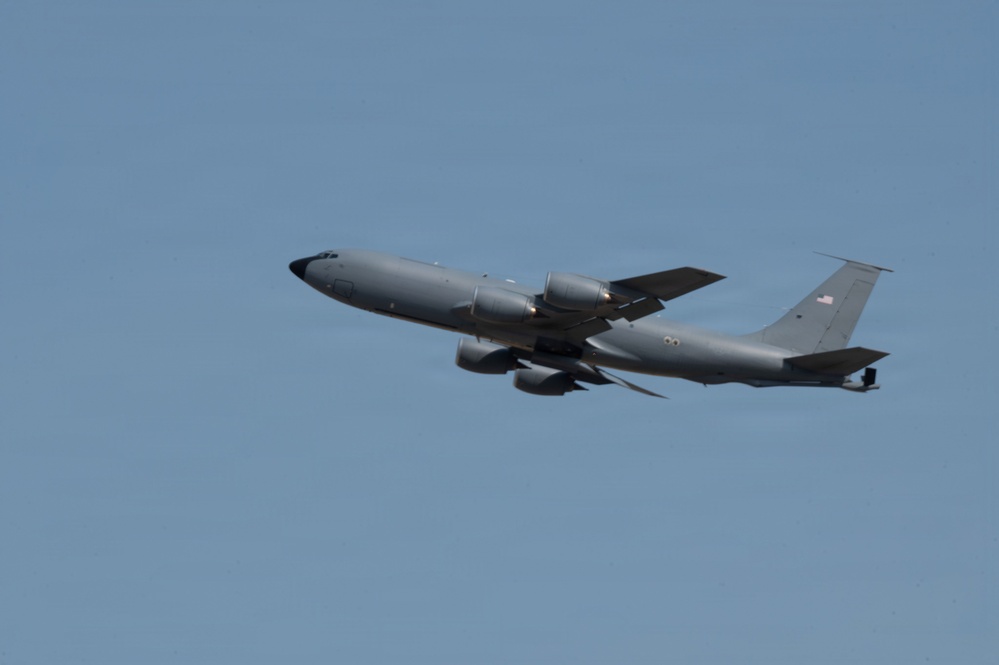 A gray military tanker aircraft, consistent with a KC-135 Stratotanker, is photographed in flight against a clear blue sky. The aircraft is in a slight climbing attitude, viewed from below and slightly to the side. An American flag is visible on the tail fin, indicating U.S. Air Force ownership.