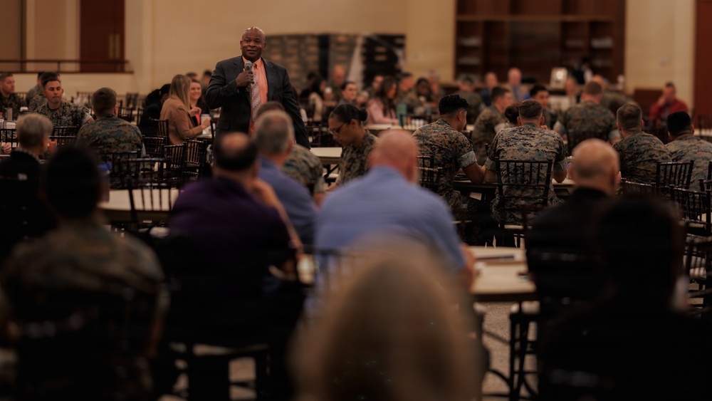 A speaker in a dark suit and orange tie addresses a large audience seated at round tables in a banquet or conference hall. The audience consists predominantly of individuals wearing military camouflage uniforms, likely U.S. Marines, along with some civilians. The setting appears to be a formal military event, briefing, or awards ceremony.