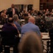 A speaker in a dark suit and orange tie addresses a large audience seated at round tables in a banquet or conference hall. The audience consists predominantly of individuals wearing military camouflage uniforms, likely U.S. Marines, along with some civilians. The setting appears to be a formal military event, briefing, or awards ceremony.