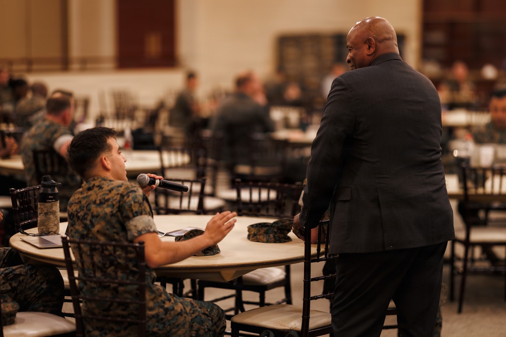 A military event or briefing taking place in a large banquet-style hall filled with personnel in camouflage uniforms. A civilian in a dark suit stands near a table while a uniformed service member holds a microphone, suggesting a Q&amp;A or presentation format. The setting appears to be a formal military gathering or leadership event.