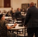 A military event or briefing taking place in a large banquet-style hall filled with personnel in camouflage uniforms. A civilian in a dark suit stands near a table while a uniformed service member holds a microphone, suggesting a Q&amp;A or presentation format. The setting appears to be a formal military gathering or leadership event.
