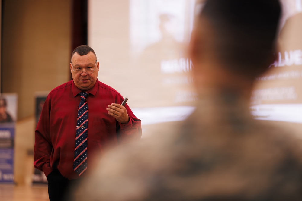 A middle-aged man in a red dress shirt and striped tie is presenting or speaking to an audience in what appears to be a formal briefing or lecture setting. He is holding a small device, likely a presentation clicker or pen, and appears to be making eye contact with his audience. A projection screen with partially visible text and imagery is visible in the background.