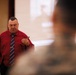 A middle-aged man in a red dress shirt and striped tie is presenting or speaking to an audience in what appears to be a formal briefing or lecture setting. He is holding a small device, likely a presentation clicker or pen, and appears to be making eye contact with his audience. A projection screen with partially visible text and imagery is visible in the background.