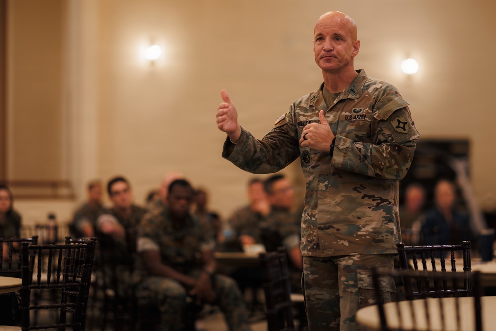 A U.S. Army officer in OCP (Operational Camouflage Pattern) uniform is addressing a group of soldiers in what appears to be a ballroom or conference hall setting. The speaker is gesturing with both thumbs up while speaking to an audience of uniformed military personnel seated at round tables. The setting suggests a formal military briefing, town hall, or leadership event.