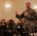 A U.S. Army officer in OCP (Operational Camouflage Pattern) uniform is addressing a group of soldiers in what appears to be a ballroom or conference hall setting. The speaker is gesturing with both thumbs up while speaking to an audience of uniformed military personnel seated at round tables. The setting suggests a formal military briefing, town hall, or leadership event.