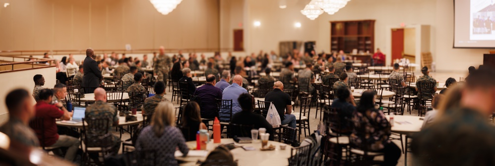 A large conference or briefing session is taking place in a banquet-style hall with numerous round tables. The audience consists of a mix of military personnel in camouflage uniforms and civilians seated throughout the room. A speaker in a dark suit is visible at the front left of the room, addressing the assembled group.