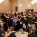 A large conference or briefing session is taking place in a banquet-style hall with numerous round tables. The audience consists of a mix of military personnel in camouflage uniforms and civilians seated throughout the room. A speaker in a dark suit is visible at the front left of the room, addressing the assembled group.