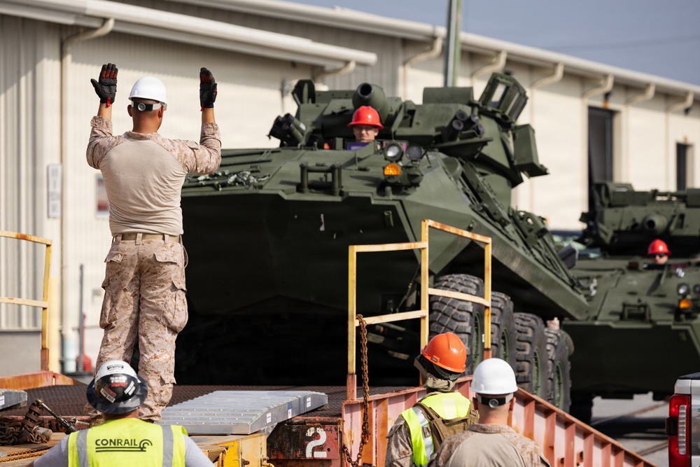 Military personnel and civilian workers are loading or positioning armored wheeled vehicles onto flatbed rail cars at what appears to be a military logistics or rail yard facility. A Marine in desert camouflage uniform is directing the movement of the vehicles with raised hands, while workers wearing safety helmets and high-visibility vests assist. At least two large green armored vehicles, consistent with LAV or Stryker-type wheeled armored personnel carriers, are visible on the rail cars.