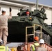 Military personnel and civilian workers are loading or positioning armored wheeled vehicles onto flatbed rail cars at what appears to be a military logistics or rail yard facility. A Marine in desert camouflage uniform is directing the movement of the vehicles with raised hands, while workers wearing safety helmets and high-visibility vests assist. At least two large green armored vehicles, consistent with LAV or Stryker-type wheeled armored personnel carriers, are visible on the rail cars.