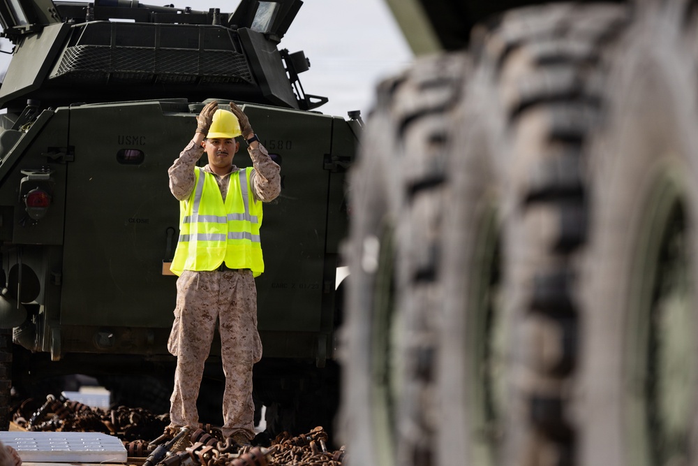 A U.S. Marine in desert camouflage uniform wearing a yellow high-visibility vest and hard hat is directing vehicle movement, with hands raised in a signaling gesture. The Marine is standing between military vehicles marked with 'USMC' markings, surrounded by chains and tie-down equipment on what appears to be a rail car or loading platform. Large military vehicle tires are visible in the foreground in soft focus.