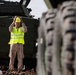 A U.S. Marine in desert camouflage uniform wearing a yellow high-visibility vest and hard hat is directing vehicle movement, with hands raised in a signaling gesture. The Marine is standing between military vehicles marked with 'USMC' markings, surrounded by chains and tie-down equipment on what appears to be a rail car or loading platform. Large military vehicle tires are visible in the foreground in soft focus.