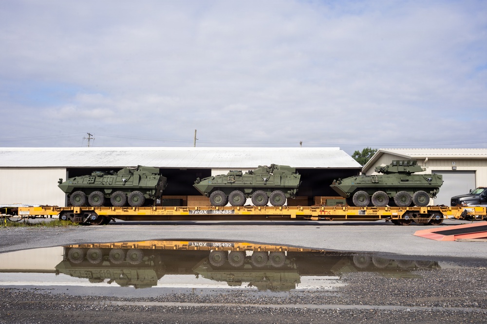 Three olive-green military wheeled armored vehicles, consistent with Stryker-type infantry carrier vehicles, are loaded on yellow railroad flatcars marked 'TPDX 192435'. The vehicles are positioned in front of a large industrial warehouse or loading facility. A puddle in the foreground creates a clear mirror reflection of the flatcars and vehicles.