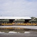 Three olive-green military wheeled armored vehicles, consistent with Stryker-type infantry carrier vehicles, are loaded on yellow railroad flatcars marked 'TPDX 192435'. The vehicles are positioned in front of a large industrial warehouse or loading facility. A puddle in the foreground creates a clear mirror reflection of the flatcars and vehicles.