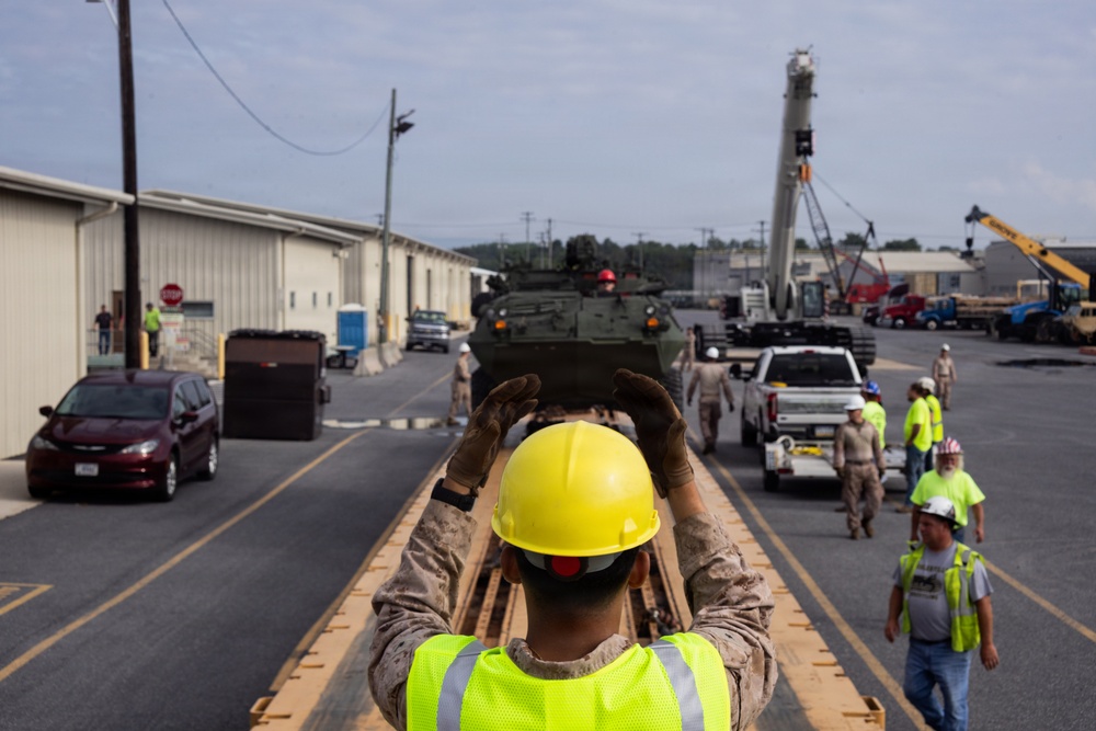 A military or industrial operation is underway in what appears to be a base or depot facility. A person wearing a yellow hard hat and high-visibility vest is directing a green armored military vehicle (likely a Stryker or similar wheeled armored personnel carrier) along a wooden ramp or track. Multiple workers in safety gear and a crane are visible in the background, suggesting a vehicle loading or positioning operation.